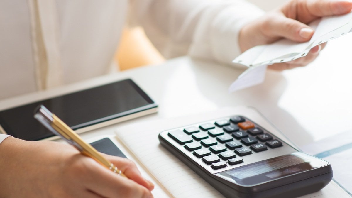 Closeup of person holding bills and calculating them. Notebook, calculator and smartphone lying on desk. Payment concept. Cropped view.
