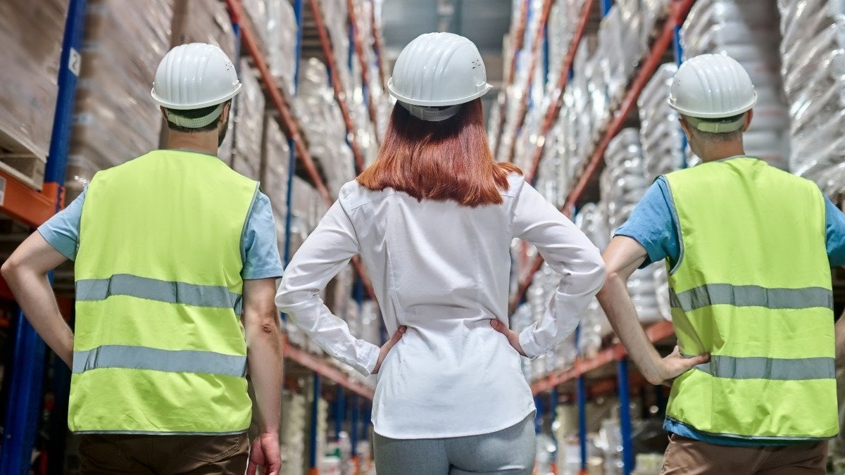 Team. Back view of long-haired female leader in protective helmet and two men in overalls standing with hands on belt in warehouse aisle