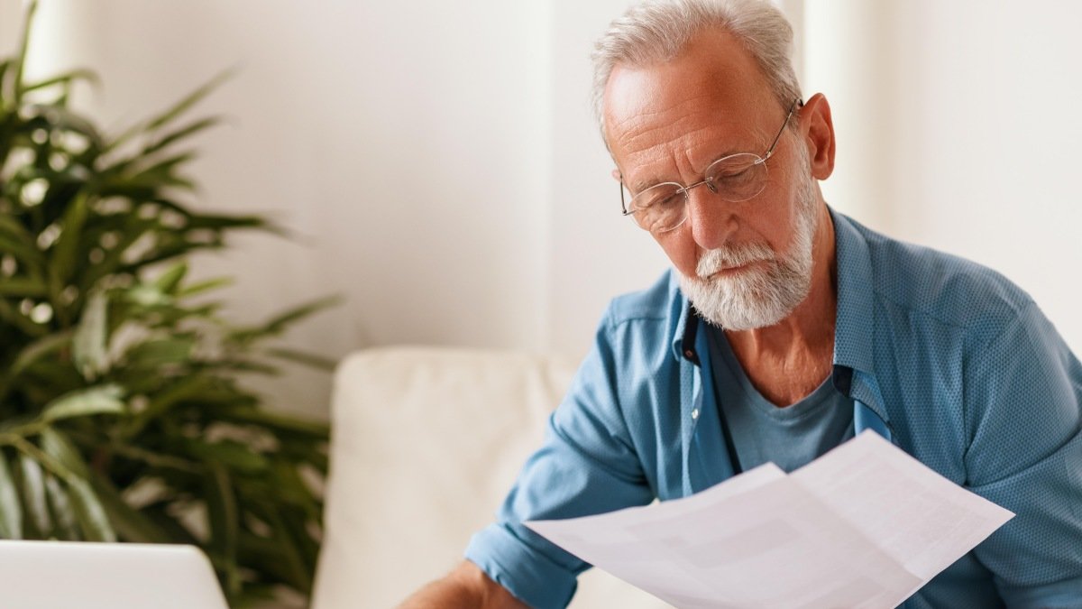 Elderly serious man in glasses reading document and making notes in laptop while sitting at table and working on freelance project at home