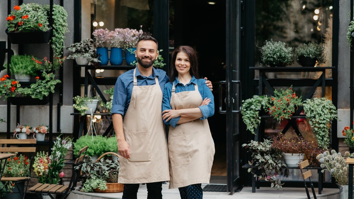 Eco cafe outdoor, family small business and client meeting after covid-19. Happy millennial male hugs female in apron with crossed arms stand near flowers shop with different plants in pots, panorama