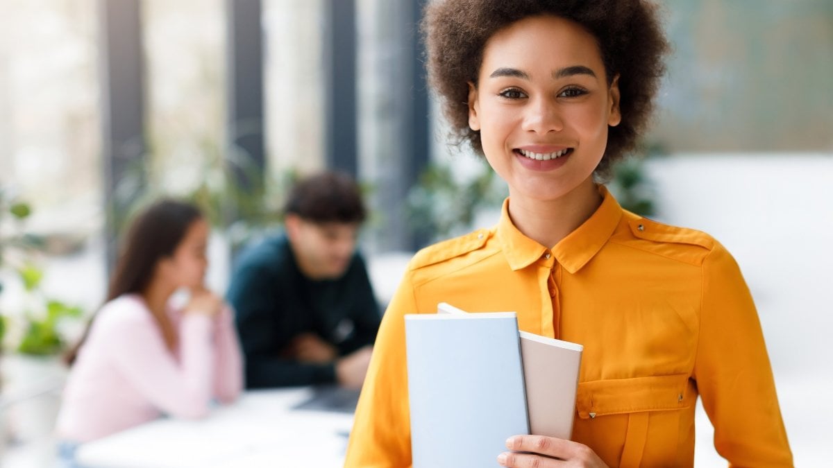 Portrait of happy black lady university student posing with notepads and smiling while her classmates studying on background in modern audience interior
