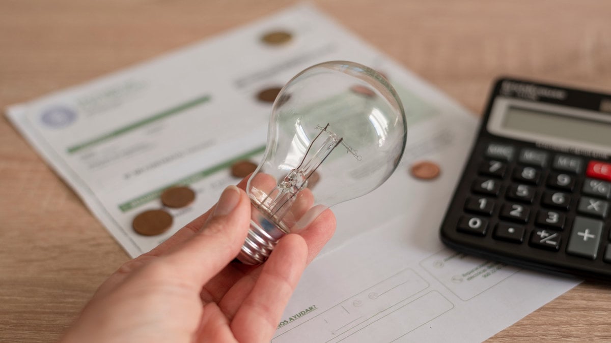 One hand holding light bulb over electricity bill, several coins and a calculator on the bill. Concept of electricity prices and tax payment.