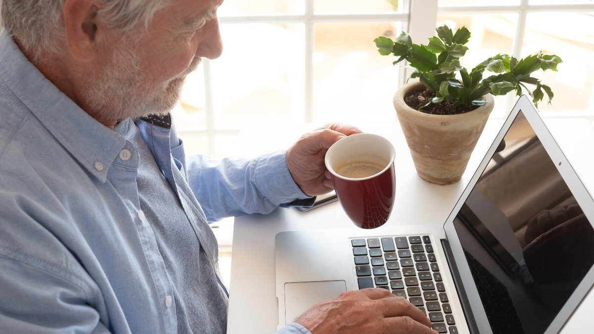 Mature senior bearded man using laptop typing on keyboard, smart work concept. Elderly white haired man sitting at desk in front the window using technology