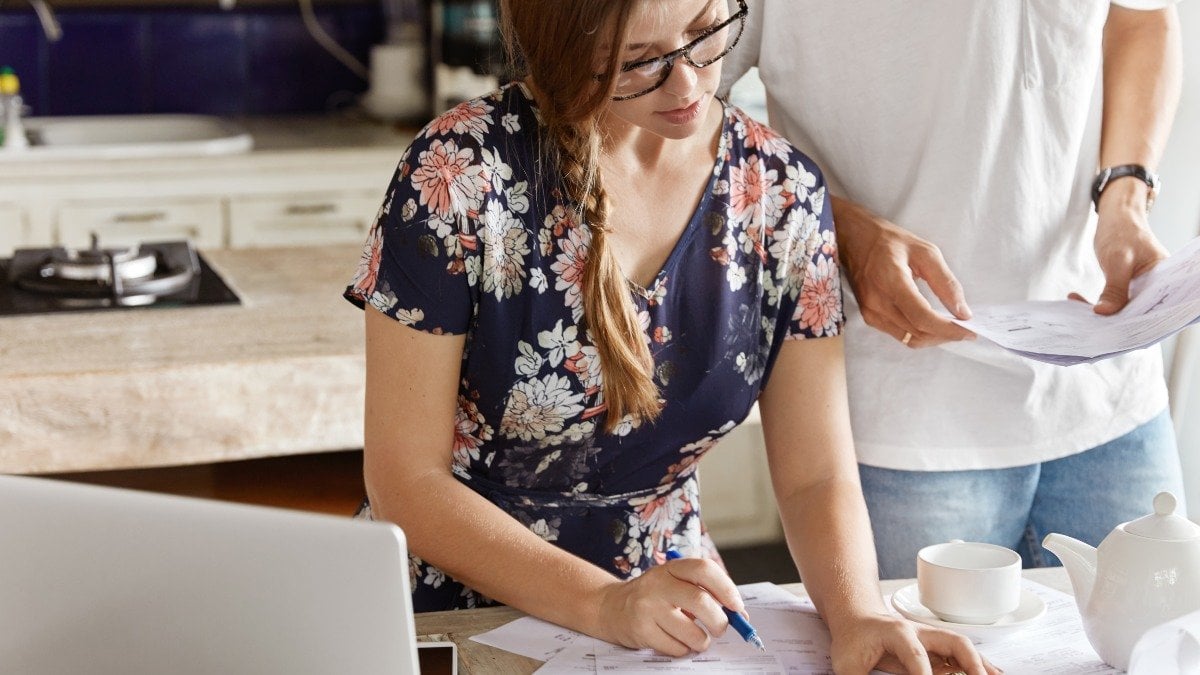 Cropped shot of pretty young woman calculates figures and writes down in papers, unrecognizable man stands near, helps with financial report. Domestic budget, family economy and expenses concept