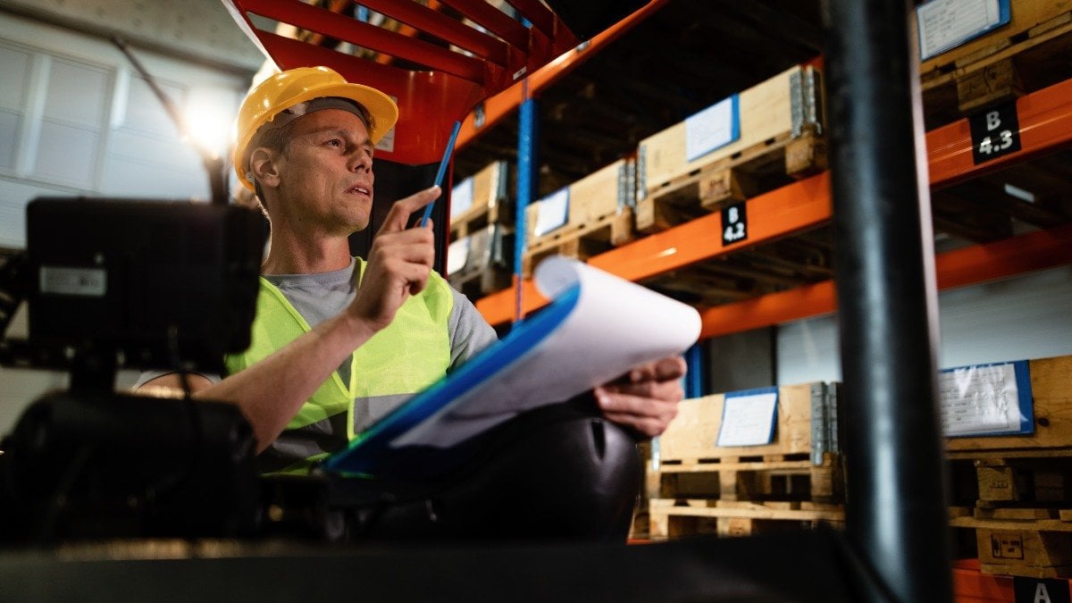 Pensive forklift operator examining stock of packages on shelves in a warehouse. 
