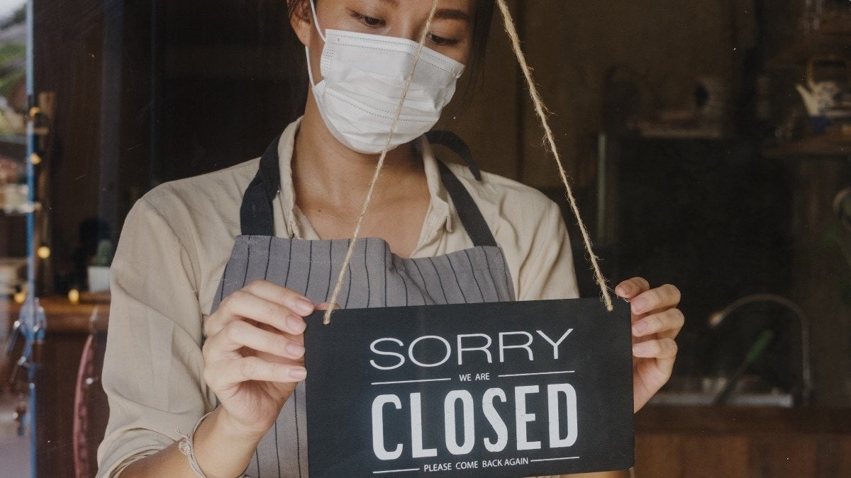 Young Asia girl wear face mask turning a sign from open to closed sign on glass door cafe after coronavirus lockdown quarantine. Owner small business, food and drink, business financial crisis concept