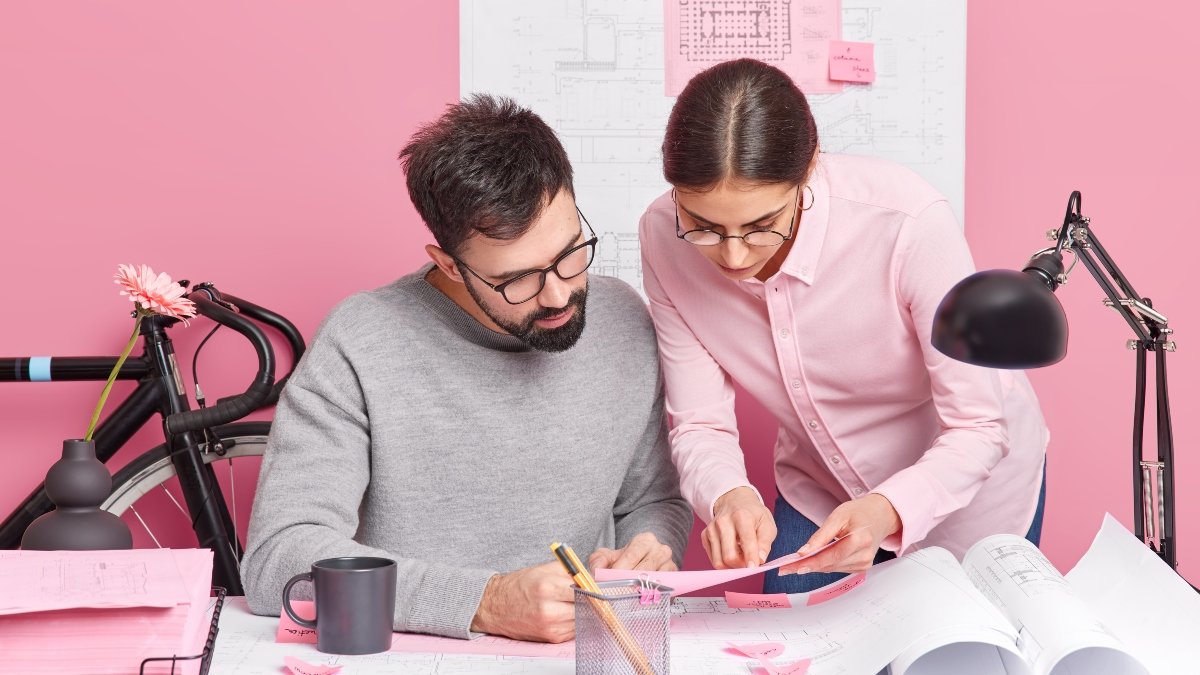 Indoor shot of busy woman and man work together on design project pose in coworking space. Two architects discuss new house plans sit at office desk with blueprints around. Collaboration concept