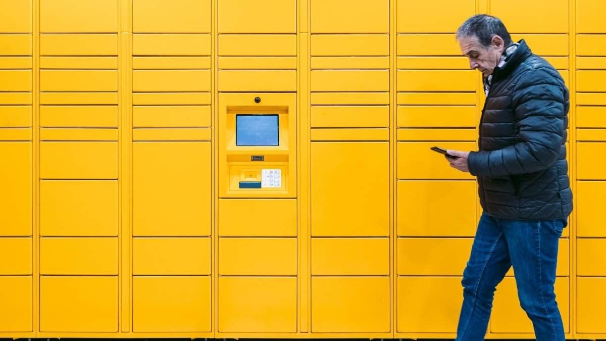 Man with gray coat looks at his cell phone to pick up a package from the locker. Messaging concept, compare online, e-commerce and packages
