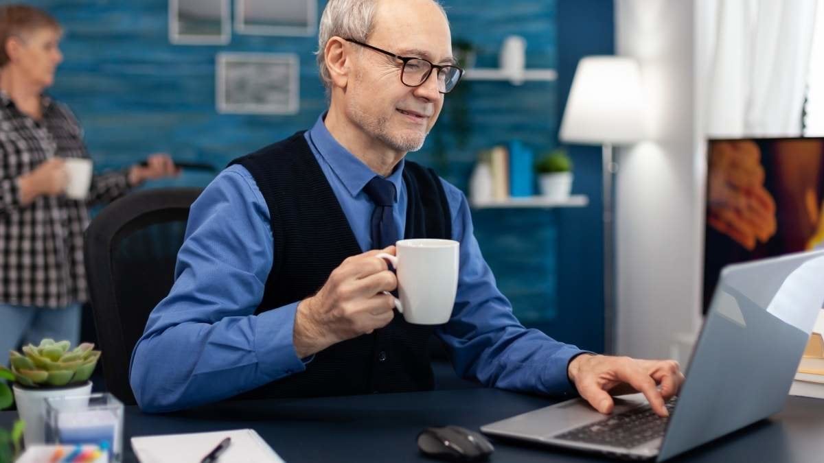 Senior businessman holding cup of coffee working on laptop. Elderly man entrepreneur in home workplace using portable computer sitting at desk while wife is holding tv remote.