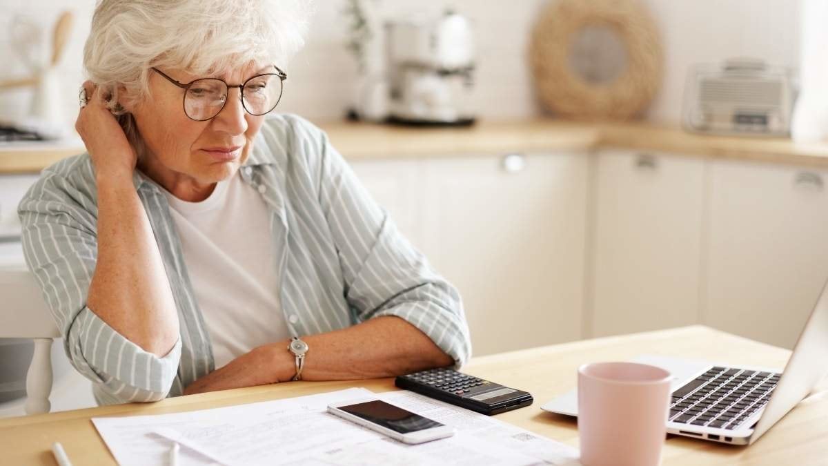 People, age, technology and finances. Depressed unhappy retired woman paying domestic bills online, trying hard to make both ends meet, sitting at kitchen table, surrounded with papers, using gadgets