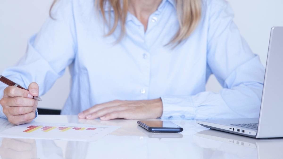 Blonde woman in blue shirt working with mobile and her laptop and writing down the results with her pen