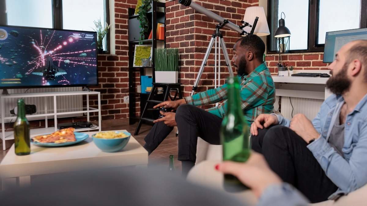 Young adult using joystick and console to play video games at home gathering with friends, drinking beer and eating snacks. Group of people having fun with gaming competition, entertainment.