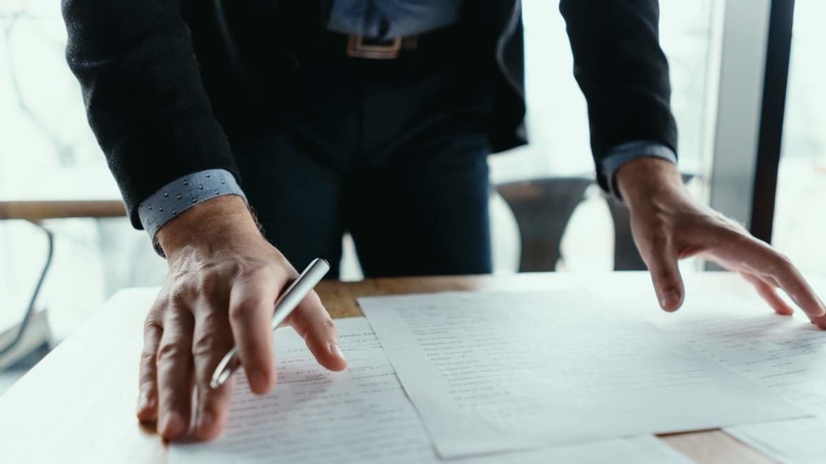 Close up hands signing documents in a modern office with window in background. Pen in hand, papers on the wooden desk, futuristic background.