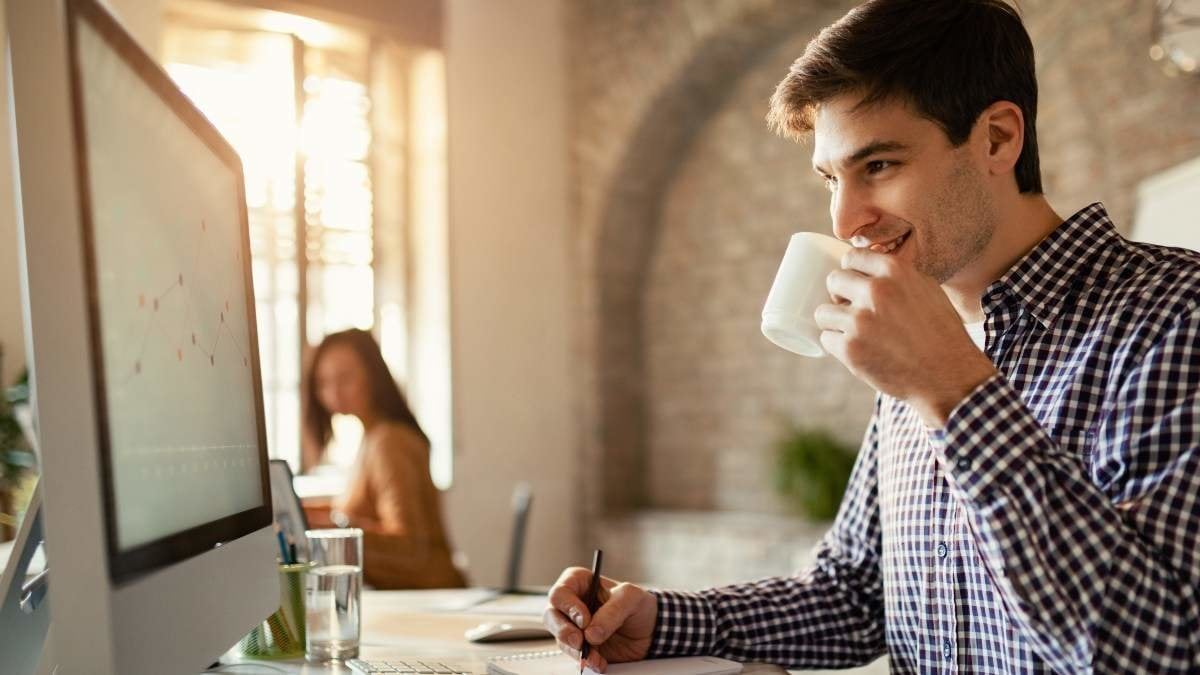 Smiling businessman drinking coffee while taking notes and using desktop PC in the office.