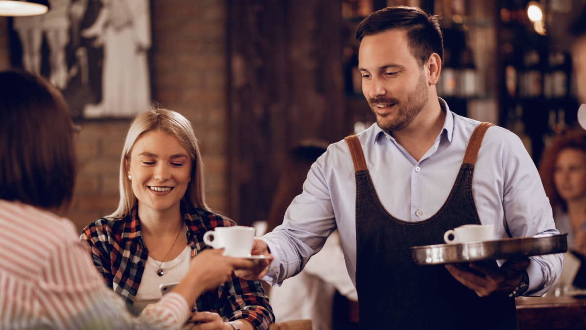 Smiling waiter serving coffee to female guests in a cafe.