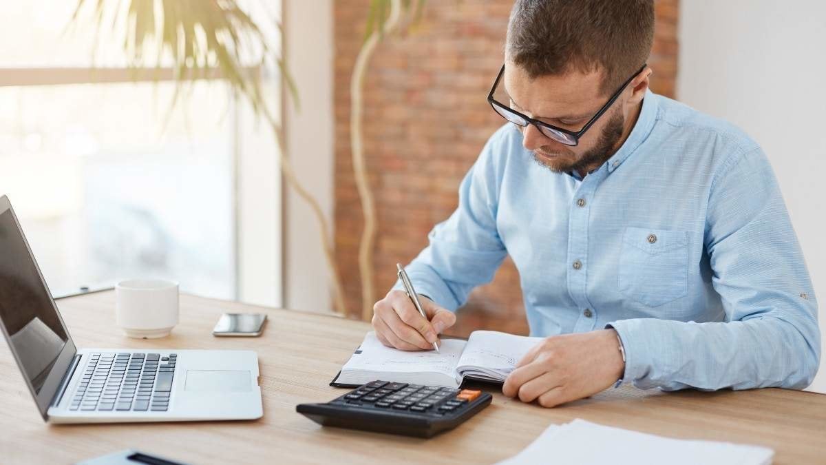 Business concept. Young unshaved caucasian male manager in glasses sitting in company office, writing down dates of meeting with customers in notebook with concentrated expression