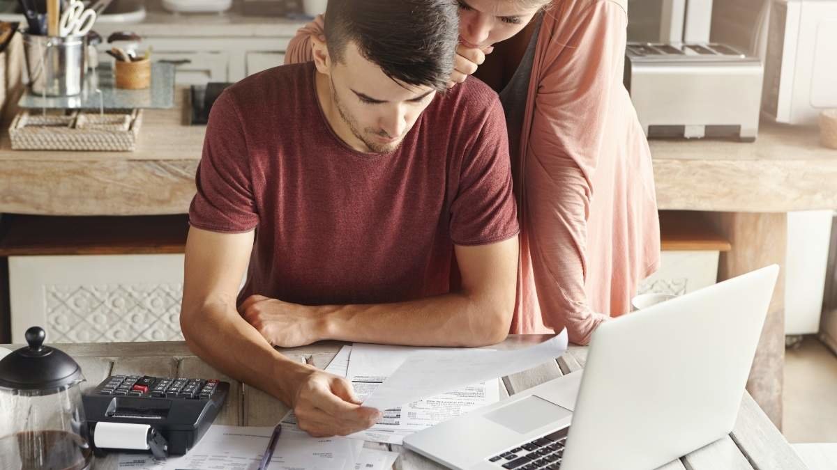 Young family managing budget, reviewing their bank accounts using generic laptop pc and calculator in kitchen. Husband and wife doing paperwork together, paying taxes online on notebook computer