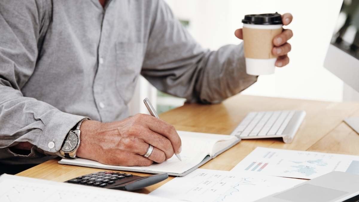 Close-up of businessman writing plans in his notepad at desk and holding cup of coffee in his hand