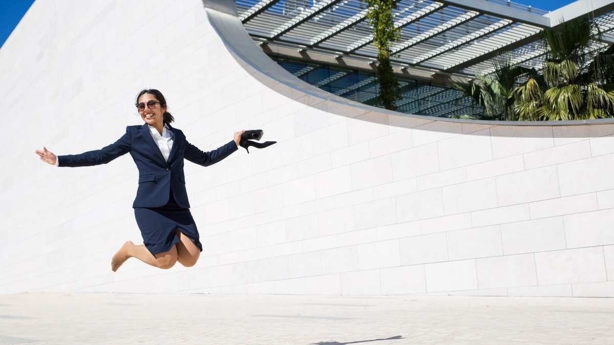 Joyful excited businesswoman celebrating success. Positive young woman in formal suit holding shoes and jumping high outdoors. Business success concept