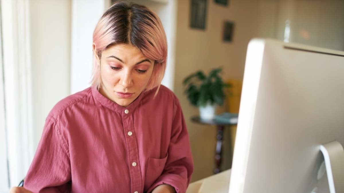 Portrait of serious young businesswoman with pink eyeshadow and hair working from home, studying financial documents, sitting at table using computer. Technology, business and occupation concept