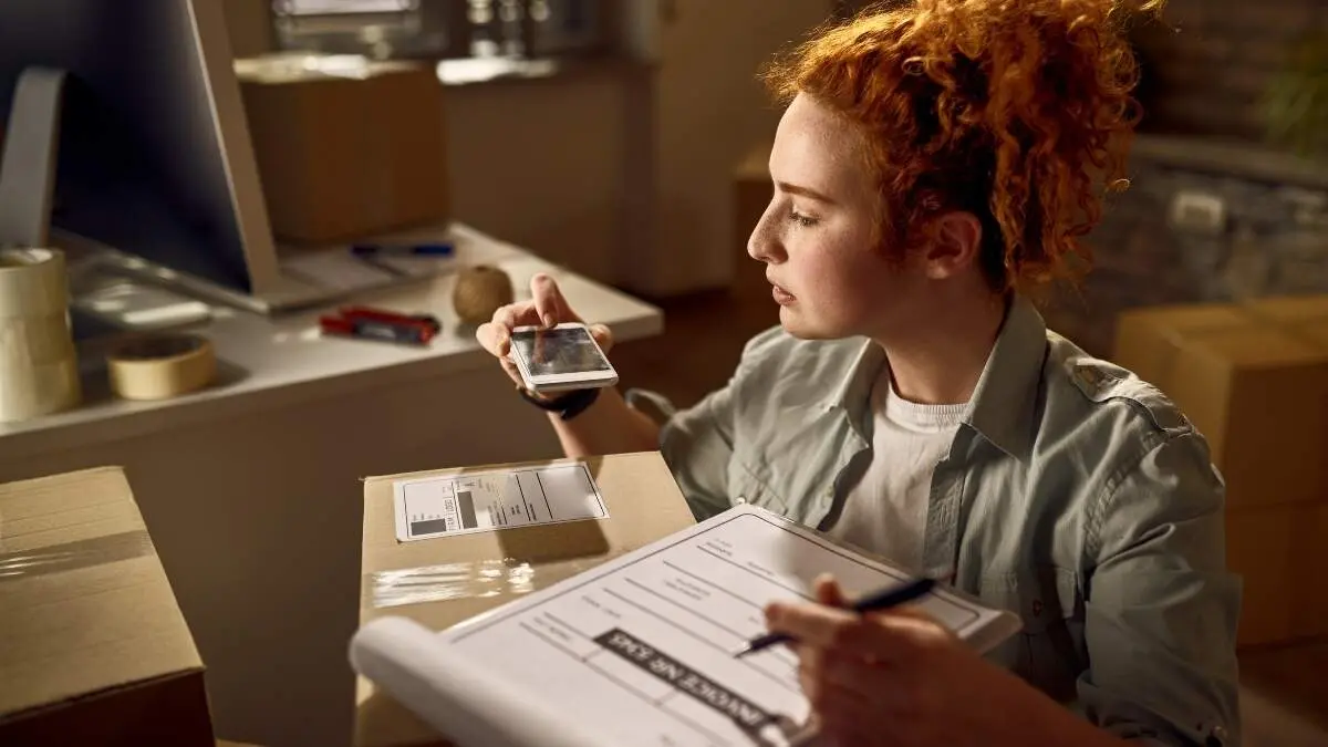 Young delivery woman using mobile phone for scanning bar code on package label while working in the office.