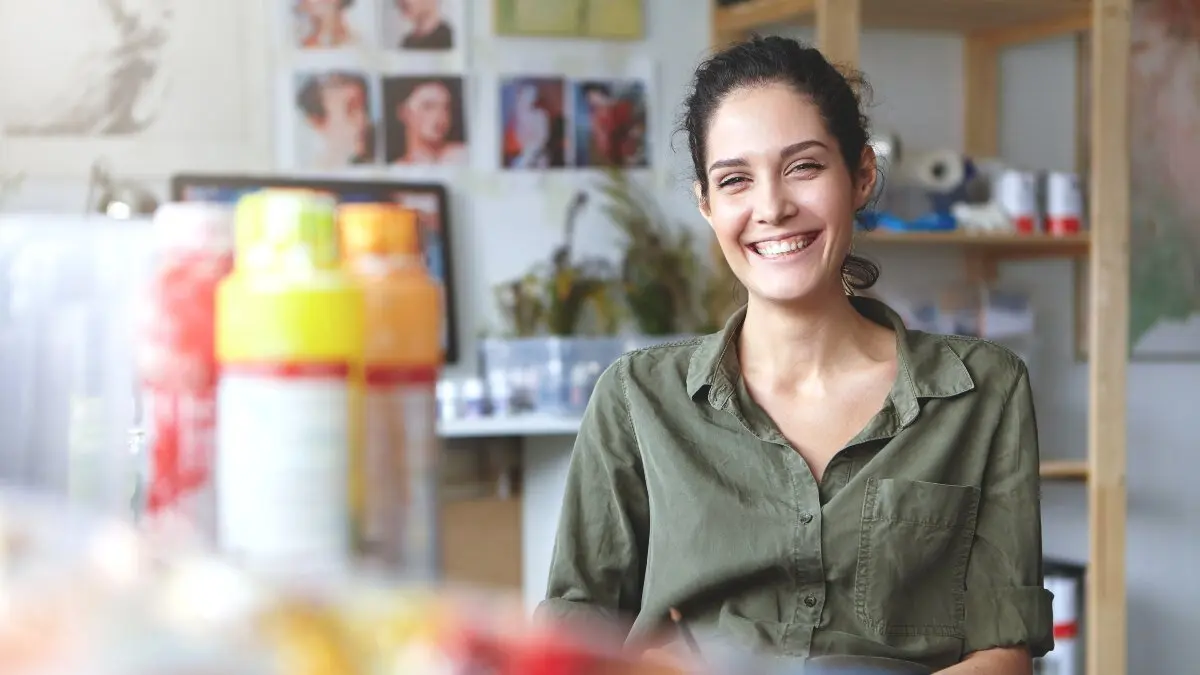 Picture of charming charismatic young female artist wearing khaki shirt grinning broadly feeling happy about her job and creating process, sitting at workshop, surrounded with painting accessories