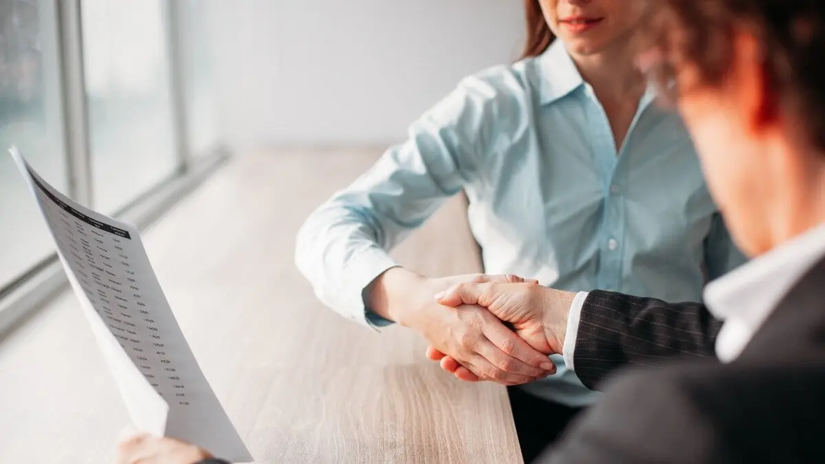 Unrecognizable businesswoman holding document and shaking hand of business partner after signing of contract. They sitting at table. Negotiation concept