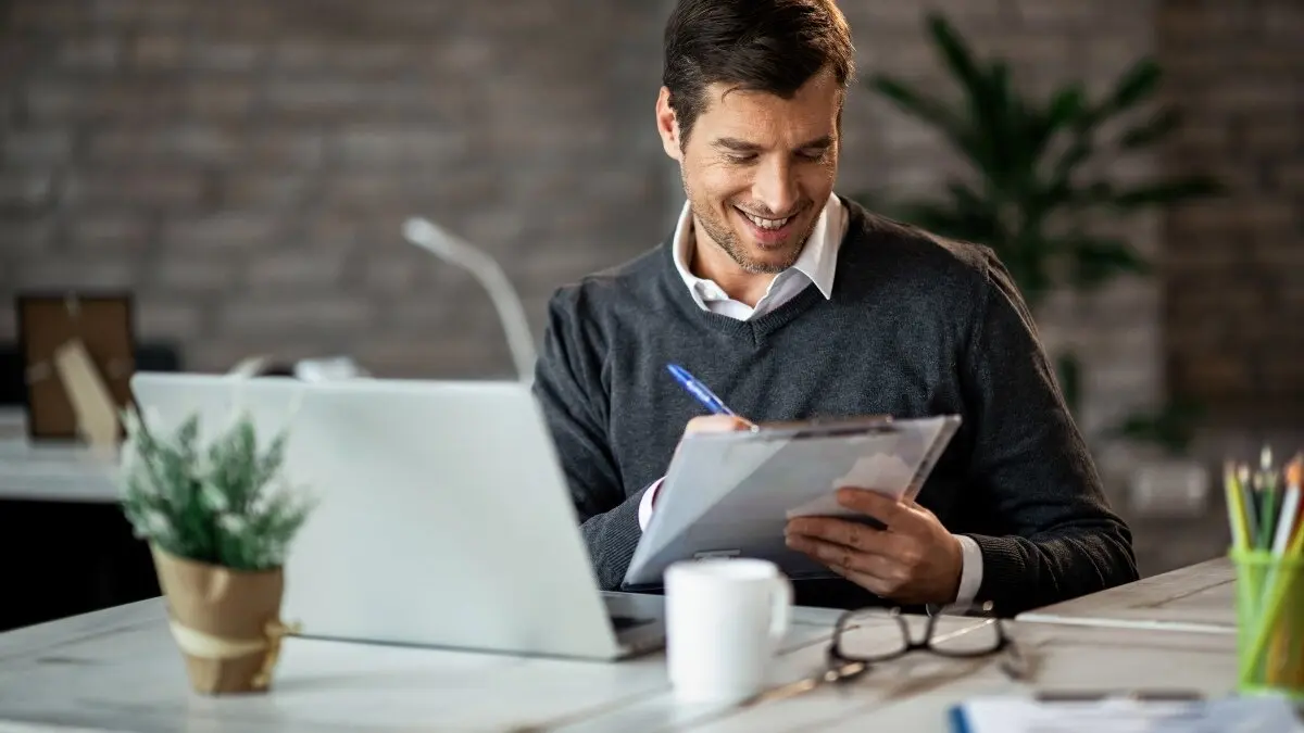 Smiling entrepreneur writing business report while working on laptop in the office.