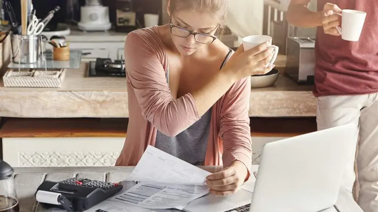 Concentrated woman dressed casually calculating bills, sitting at kitchen table with laptop, calculator, papers and mobile, holding white cup and passing it to her husband, who is standing behind her