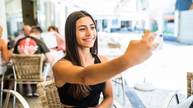 Portrait of young latin girl taking selfie with mobile phone while sitting at a cafe outdoors