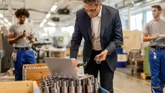 Factory manager reading instructions on a computer while checking measurements of steel bolt in industrial building. Manual workers are in the background.