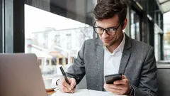 Smiling business man in eyeglasses sitting by the table in cafe with laptop computer while using smartphone and writing something