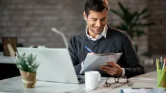 Smiling entrepreneur writing business report while working on laptop in the office.