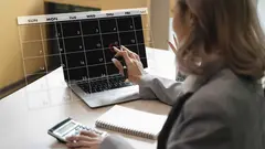 A 2025 calendar woman business suit is planning her schedule digital calendar overlay, using pen and laptop, with calculator and notebook nearby