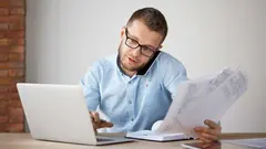 Business concept. Multitasking. Serious professional good-looking businessman talking on phone with customer, holding papers in hand, looking for information on laptop computer