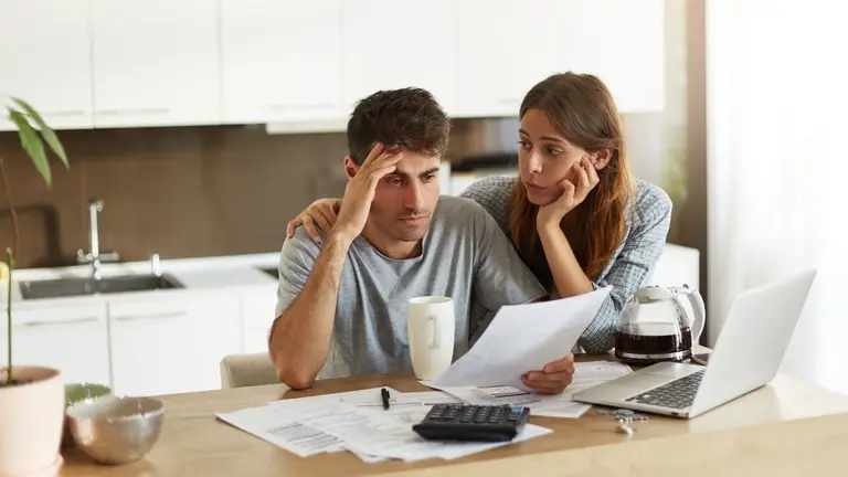 Depressed young Caucasian couple calculating family budget in kitchen, trying to find a way to make both ends meet, having stressed and frustrated looks: caring wife embracing her unhappy husband